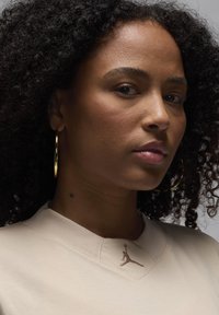 Felpa beige a girocollo con scollo strutturato e logo Jumpman ricamato in oro; il modello indossa grandi orecchini a cerchio dorati e ha i capelli ricci.