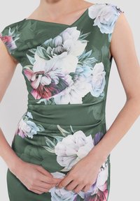 Blomsterkjole i grøn stof med store hvide og lyserøde blomster, tætsiddende design, asymmetrisk halsudskæring og rynket tekstur ved taljen.