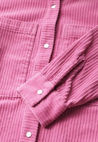 Rosa flanellskjorta med vertikal ribbning, två framfickor och metallknappdetaljer vid knäppningen och ärmsluten. Mjuk textur.