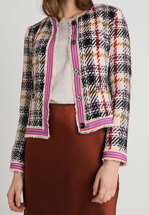 Frau trägt eine mehrfarbige karierte Jacke mit Knöpfen und Fransenbesatz über einem beigen Oberteil und einem braunen Rock, steht vor einem schlichten Hintergrund.