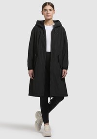 Zwarte lange jas van lichtgewicht stof, met een capuchon, ritsluiting en twee zijzakken. Model heeft het gecombineerd met zwarte leggings en sneakers.