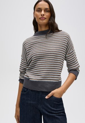 Donna che indossa un maglione a righe grigio, marrone e bianco con maniche a tre quarti e jeans blu scuro, in piedi con una mano in tasca.