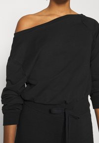 Sudadera negra con hombros descubiertos, mangas largas, puños acanalados y un material de vellón texturizado. Cuenta con una cintura con cordón ajustable.