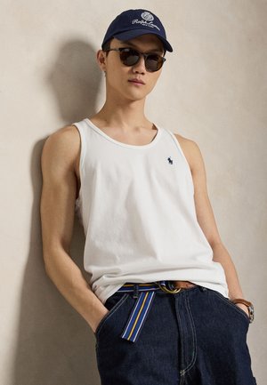 Tânăr purtând tricou alb fără mâneci, blugi închiși la culoare cu curea cu dungi, șapcă bleumarin, ochelari de soare și ceas de mână, sprijinit de un perete bej.