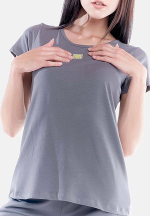 Mujer con camiseta gris con el logo "LEONE" en el pecho, sosteniendo la camiseta cerca del logo con ambas manos sobre fondo blanco.