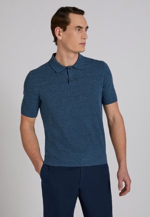 Uomo che indossa una polo a maniche corte aderente in maglia blu e pantaloni blu scuro, in piedi con una mano in tasca, che guarda di lato.