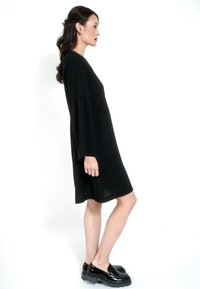 Crna haljina A kroja s dugim širokim rukavima. Glatka tekstura s labavim krojem, nosi se uz sjajne crne cipele na potpeticu s debelim đonom.