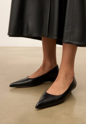 Vrouw draagt zwarte leren ballerina's met puntige neus, gecombineerd met een zwarte leren midi-rok, staand op een gladde vloer.