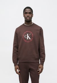 Hnedá bavlnená mikina s okrúhlym grafickým logom "Calvin Klein New York" v červenej farbe. Dizajn s okrúhlym výstrihom a rebrovanými manžetami.