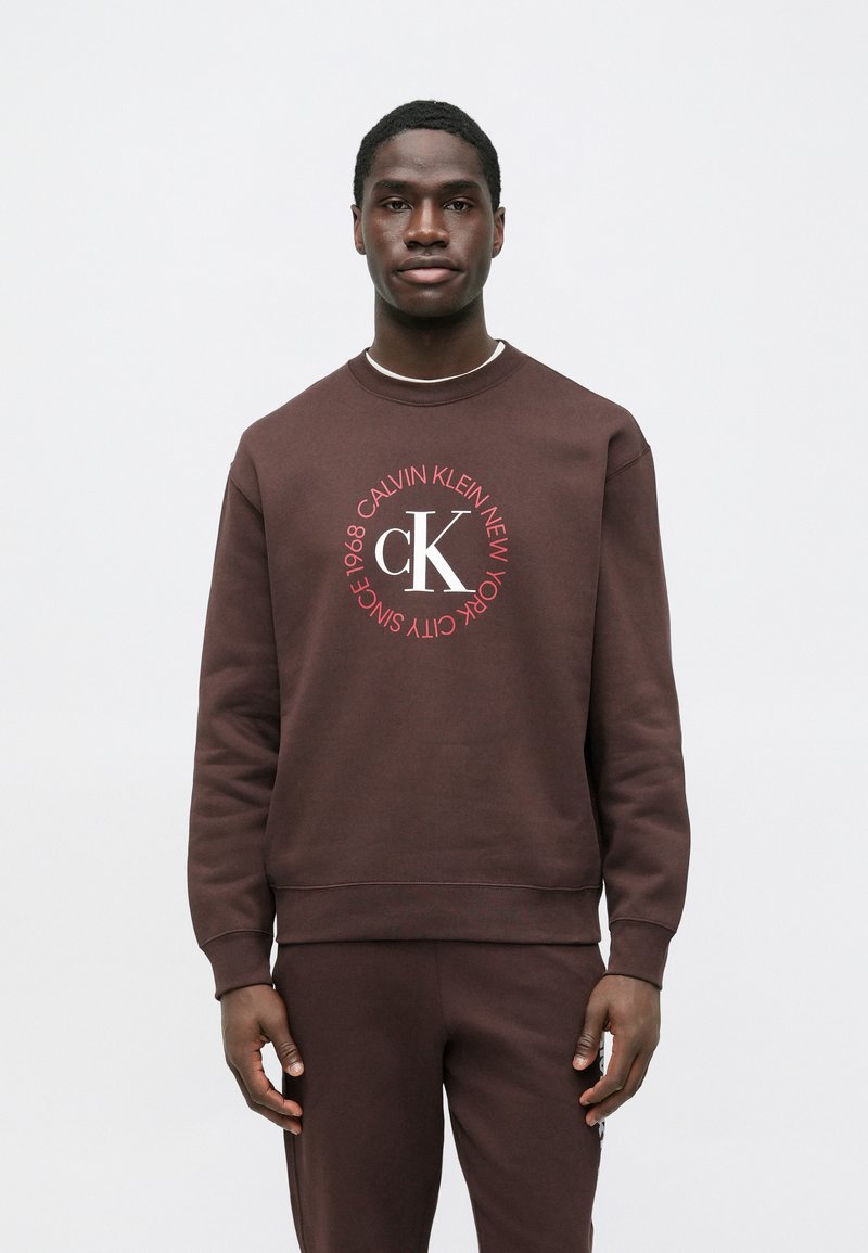 Hnedá bavlnená mikina s okrúhlym grafickým logom "Calvin Klein New York" v červenej farbe. Dizajn s okrúhlym výstrihom a rebrovanými manžetami.