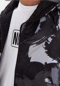 Schwarz-weiß gemusterte Jacke mit Reißverschluss, darunter ein schlichtes weißes T-Shirt mit einem grafischen Druck.