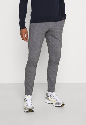 Homme portant un pantalon slim gris à carreaux, des chaussettes blanches, des baskets blanches et jaunes, et une chemise sombre à manches longues, debout devant un fond uni.