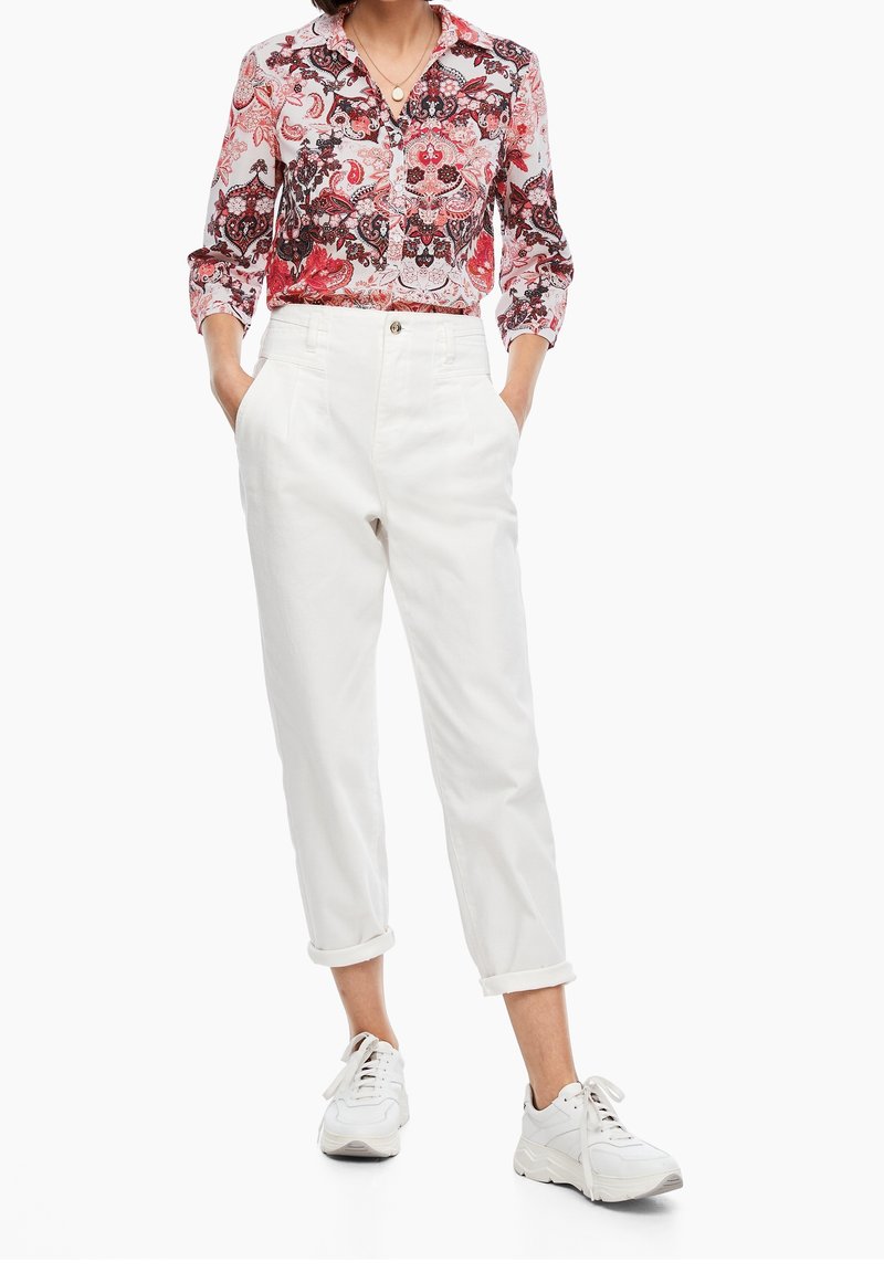 Femme portant une chemise à fleurs rouge et blanche boutonnée, un pantalon blanc taille haute retroussé et des baskets blanches, debout les mains dans les poches.