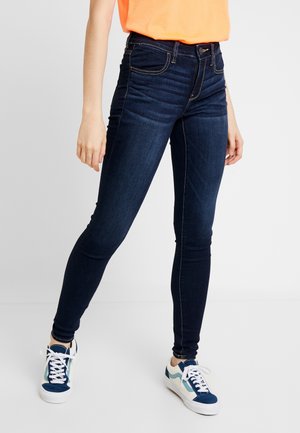 Person trägt dunkelblaue Skinny-Jeans, orangefarbenes Oberteil und weiß-blaue Sneakers, steht vor einem einfarbigen hellen Hintergrund.