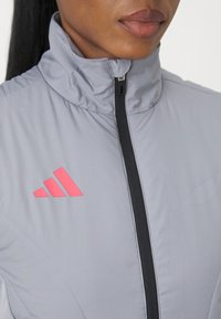 Pilkas sportinis švarkas su aukštu apykakle, pilnu zilpimo užtrauktuku ir rožinės spalvos Adidas logotipu kairėje krūtinės pusėje. Lygi tekstūra, modernus dizainas.