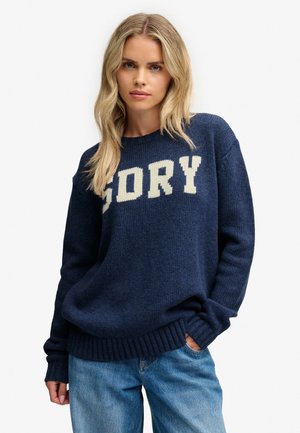 Kvinde med langt blondt hår iført en marineblå sweater med ordet "SORY" og blå jeans med den ene hånd i lommen.