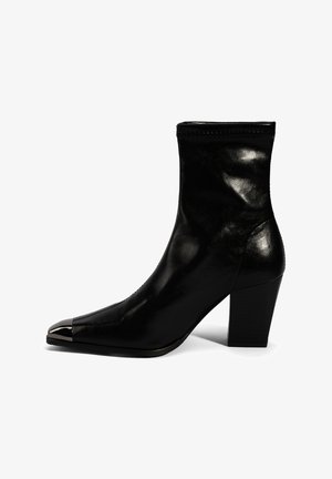 Bottine en cuir noir à bout pointu, avec un accent métallique sur le capuchon, et un talon empilé. Texture lisse et design épuré.