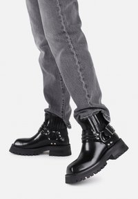 Botas de cuero negras hasta el tobillo con suela gruesa, hebillas de metal y detalle elástico en la parte superior, combinadas con pantalones grises con vuelta.