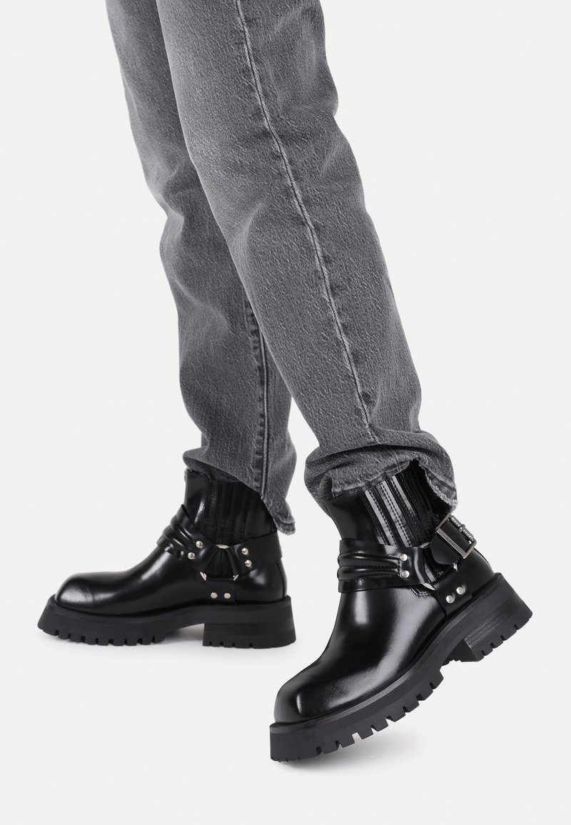 Schwarze Leder-Stiefeletten mit einer robusten Sohle, metallischen Schnallen und elastischen Details am oberen Rand, getragen mit grauen hochgekrempelten Hosen.