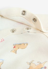 Nærbillede af en cremefarvet babytøj med metalknapper og små trykte illustrationer af hunde med røde sløjfer og blå halsbånd.