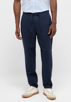 Mann trägt marineblaue Hose mit Kordelzug, kombiniert mit weißen Turnschuhen und hellblauem Hemd, steht vor einem einfarbigen Hintergrund.