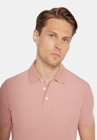 Różowa koszulka polo wykonana z dzianiny, z kołnierzykiem, trzema guzikami oraz dyskretnym znakiem logo w okolicy klatki piersiowej.