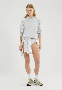 Grauer Kapuzenpullover, weiß gestreifter Wickelrock um die Taille, weiße Shorts, beige Sneaker mit grünen Akzenten und weiße Knöchelsocken.