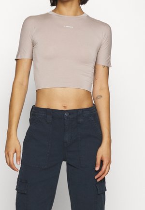 Person trägt ein hellbeiges, kurzes T-Shirt und hochtaillierte dunkle Cargo-Hosen, zeigt den Mittelteil und Arm-Tattoos.