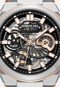 Automatyczny zegarek z czarną szkieletem tarczą, różowozłotymi akcentami i srebrną stalową kopertą. Wyposażony w skomplikowane zębatki i charakterystyczne znaczniki.