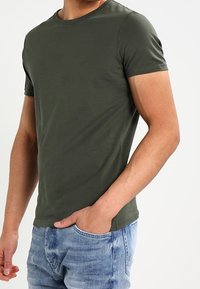 Muž oblečený v těsném olivově zeleném tričku s krátkým rukávem a světlých denimových jeansách, s jednou rukou v kapse, na neutrálním pozadí.