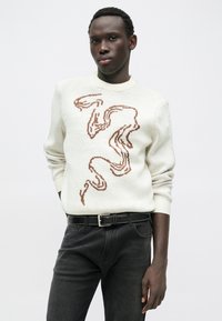 Pleteni pulover v krem barvi z rjavim abstraktnim vzorcem, okrogel izrez in rebrasti manšeti. Nošen z temnimi denim kavbojkami in črnim pasom.