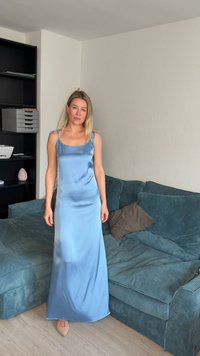 Femme aux cheveux blonds portant une robe en satin bleu clair, debout à côté d'un canapé d'angle turquoise avec des coussins dans un salon.