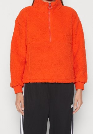 Person trägt einen leuchtend orangefarbenen Fleece-Pullover mit halbhohem Reißverschluss vorne und schwarze Adidas-Trainingshose mit weißen Streifen.