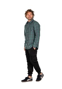 Camisa de cuadros verde con un bolsillo; pantalones cargo negros; zapatillas negras con detalles blancos. Modelo de pie con las manos en los bolsillos.