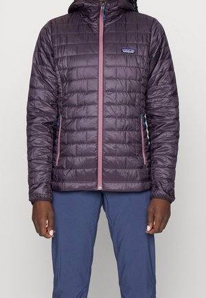 Personne portant une veste matelassée Patagonia violette avec une fermeture éclair rose et un pantalon bleu, debout devant un fond uni.