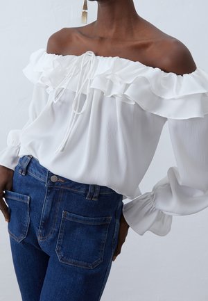 Femme portant un chemisier blanc à épaules dénudées avec des volants et des manches longues, assorti à un jean en denim bleu taille haute et des boucles d'oreilles géométriques.