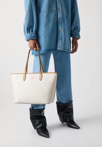 Tasche mit cremefarbenem Karomuster, hellbraunen Ledergriffen und glatter Oberfläche. Kombiniert mit einem Denim-Outfit und schwarzen spitzen Stiefeln.