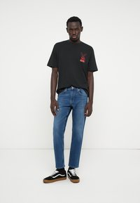 Camiseta de algodão preta com design gráfico vermelho no peito, combinada com jeans de ganga azuis e ténis pretos com pormenores brancos.