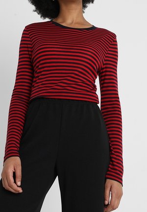 Kobieta ubrana w dopasowaną, długą bluzkę z czerwono-czarnymi poziomymi paskami, zestawioną z czarnymi spodniami z wysokim stanem.