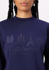 Sweatshirt bleu marine avec des monuments parisiens en relief : Notre-Dame, Sacré-Cœur, Tour Eiffel et Arc de Triomphe, avec le texte "Paris" en dessous.