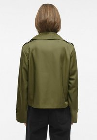 Izgriezta olīvzaļā jaka ar gludu tekstūru, izgrieztu apkakli un pogu detaļām uz piedurknēm; veidota ar gludu siluetu.