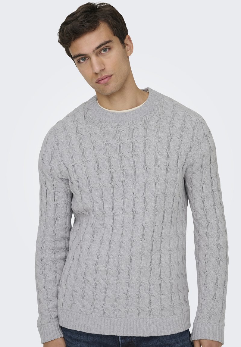 Jeune homme aux cheveux courts et foncés portant un pull gris clair en maille torsadée et un jean bleu, debout devant un fond clair uni.