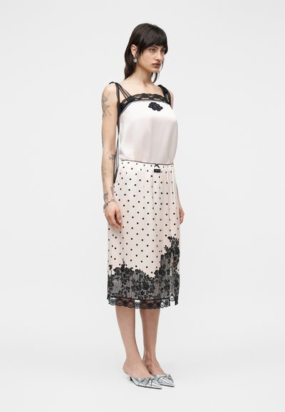 Γυναίκα με λευκό αμάνικο φόρεμα με μαύρη δαντέλα στο τελείωμα, πουά σχέδιο, floral μοτίβο στο τελείωμα, ασημί μυτερές γόβες, στέκεται μπροστά σε απλό φόντο.