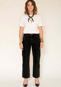 Femme se tenant contre un mur beige, portant un t-shirt blanc, un jean noir ajusté, des talons noirs, un foulard noir au cou, des bracelets en or et des bagues.