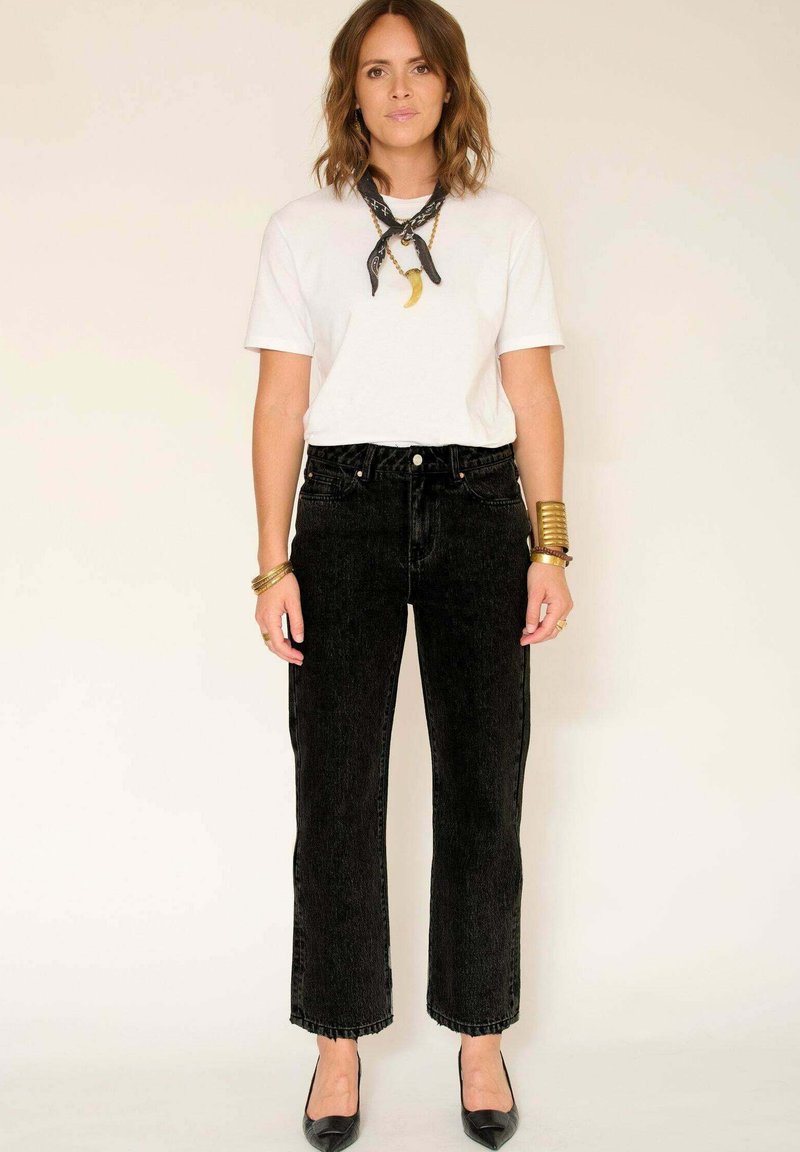 Femme se tenant contre un mur beige, portant un t-shirt blanc, un jean noir ajusté, des talons noirs, un foulard noir au cou, des bracelets en or et des bagues.