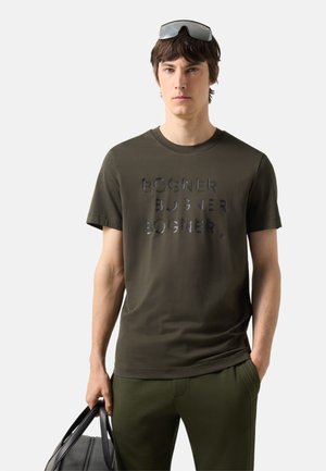 Ung mand iført olivengrøn Bogner t-shirt og bukser, der holder en sort taske, med solbriller hvilende på sit hoved, neutral baggrund.
