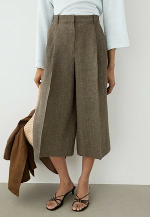 Pantalon en lin marron large et court, porté avec un haut blanc, des sandales à talons noires à brides, tenant une veste marron et un sac tressé.