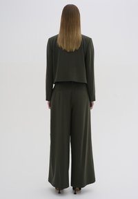 Olivgrüner, taillierter Blazer über weitem Hosen. Glattes Material mit minimalen Nähten und zwei Gesäßtaschen. Mittellange, glatte Haare.