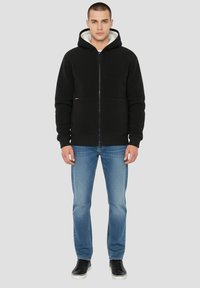 Schwarzer Hoodie mit Reißverschluss, weichem cremefarbenem Futter und zwei Vordertaschen, kombiniert mit blauen Jeans und schwarzen Sneakern. Glatte Textur und schlichtes Design.