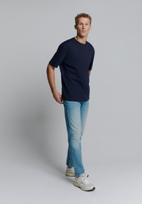 Navy blauwe t-shirt met korte mouwen gecombineerd met lichtblauwe spijkerbroek. Het model draagt witte en grijze sneakers. Effen achtergrond.